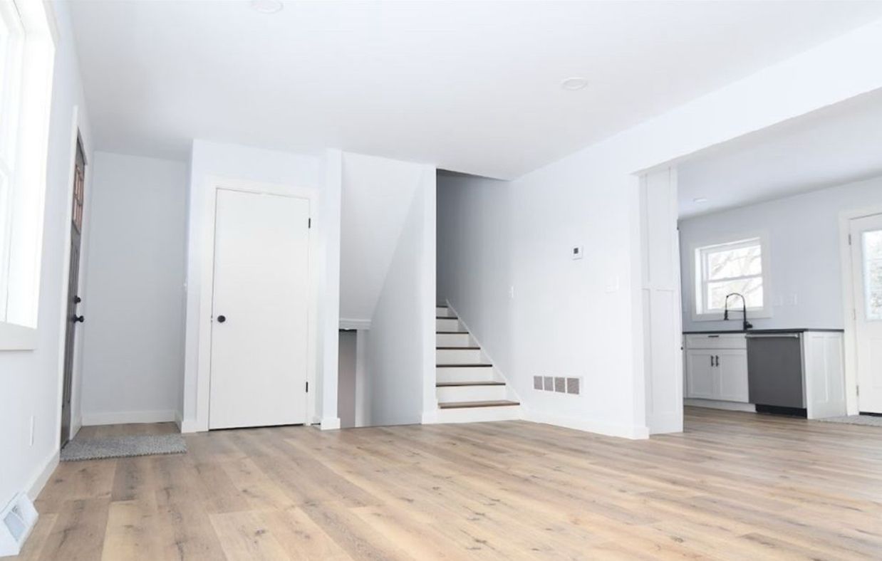 A bright, empty living room with light wood flooring, white walls, stairs, and a view into a kitchen with grey cabinets.