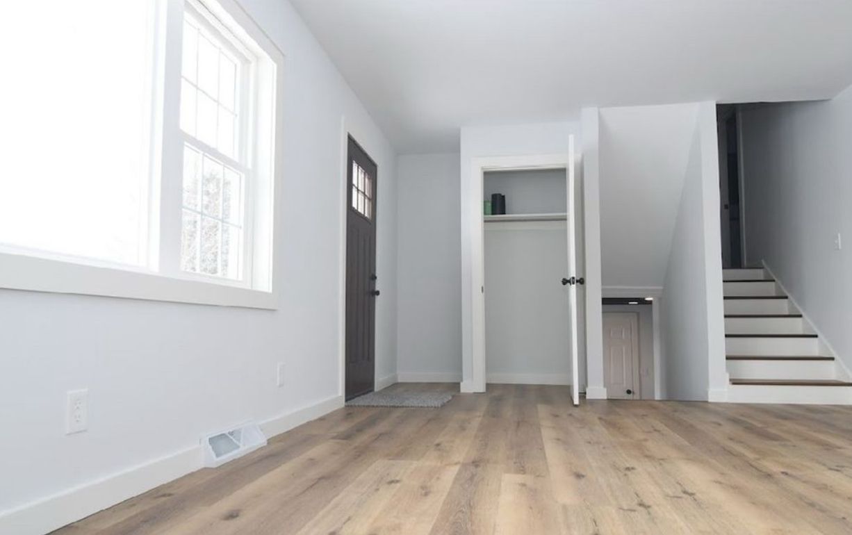 A brightly lit room with white walls, wood-look flooring, a window, a dark front door, a closet, and stairs going upstairs.