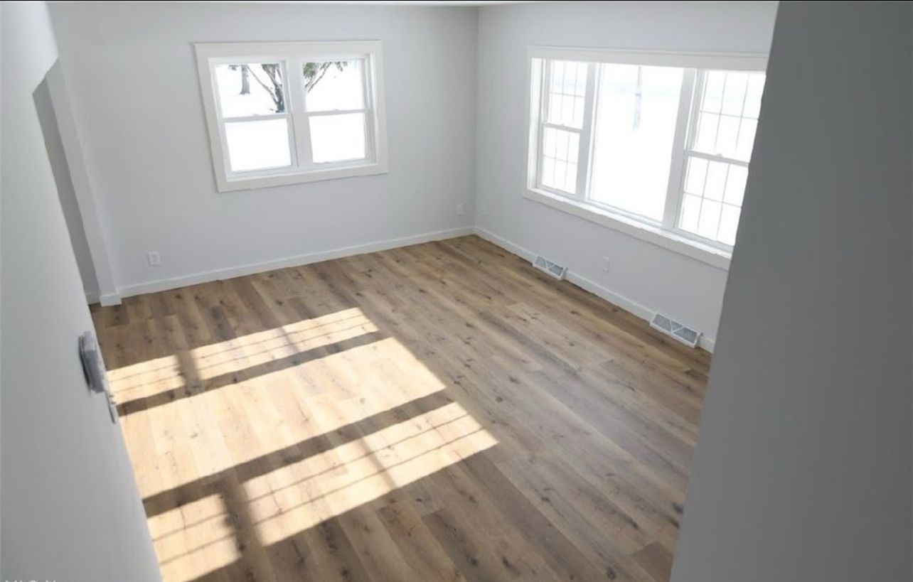 Empty room with light gray walls, wood-look flooring, and natural light streaming through two windows.