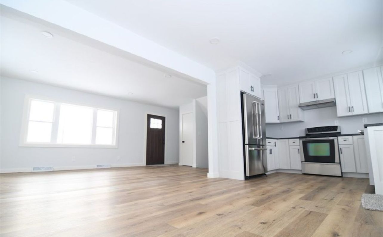 An open-concept room with light hardwood floors, white kitchen cabinets, stainless steel appliances, and a dark door.