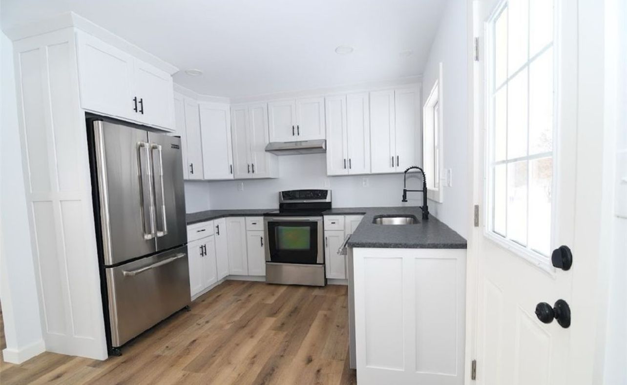 A modern kitchen with white cabinets, stainless steel appliances, dark countertops, and light wood-look flooring.