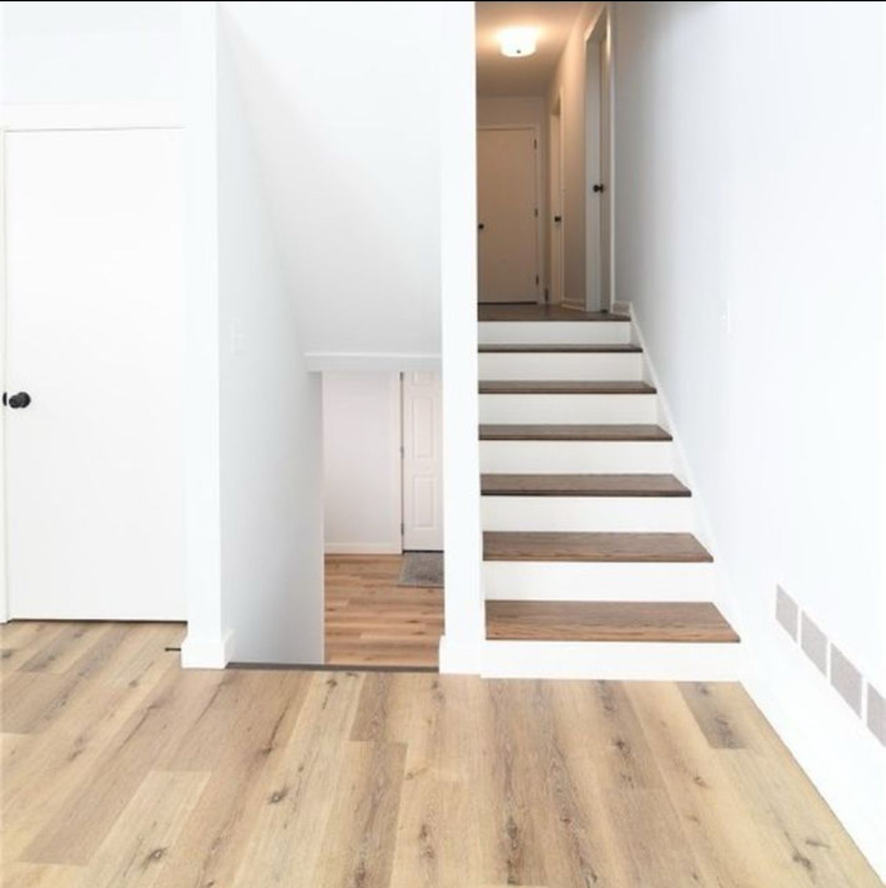 An interior view of a modern hallway featuring light wood flooring, white walls, and a staircase with wooden treads.