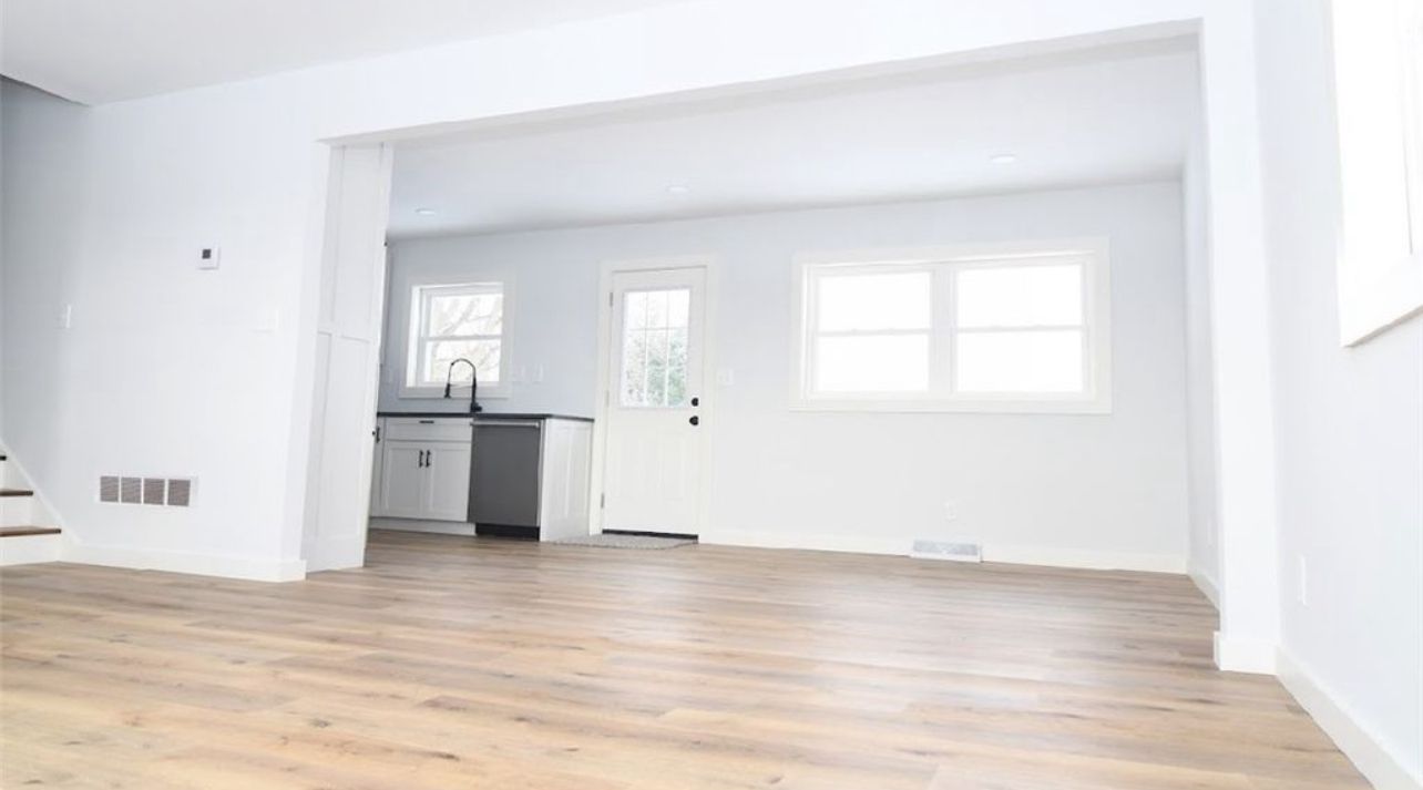 A bright, open-plan room with light wood flooring, white walls, and a glimpse into a kitchen with a white door.