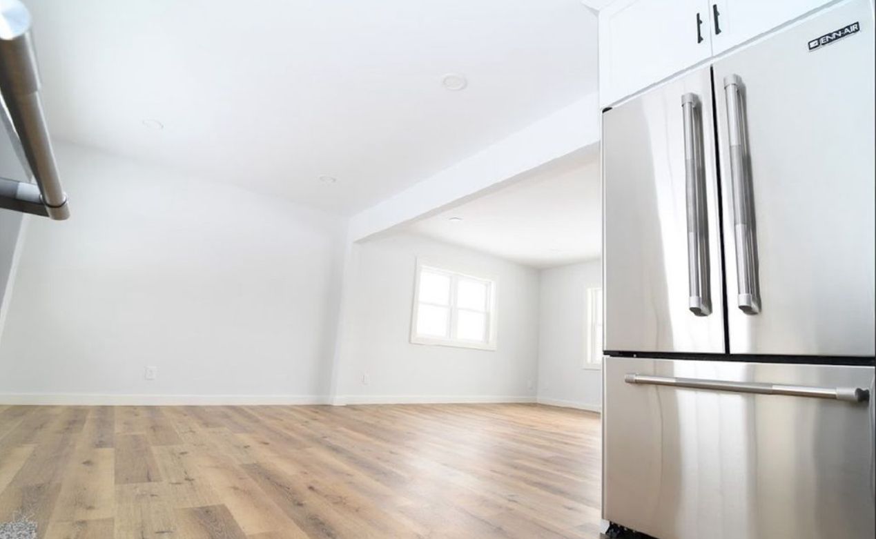 A modern kitchen with light wood-tone flooring, white walls, and a stainless steel French-door refrigerator.