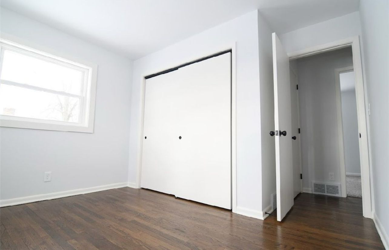 Empty room with white walls, dark wood floors, a closet with white sliding doors, and an open doorway leading to a hallway.