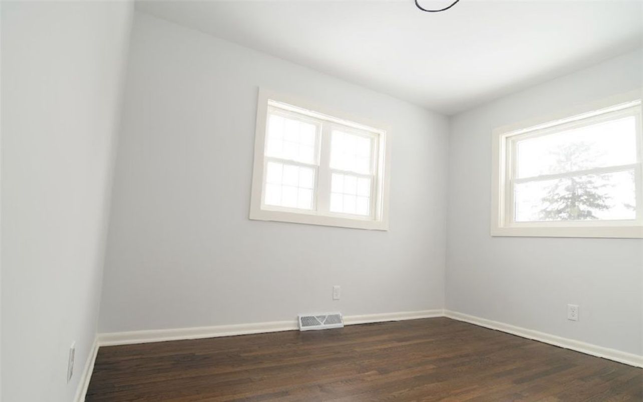 A sparsely furnished room with light gray walls, dark wood flooring, and two windows allowing natural light.