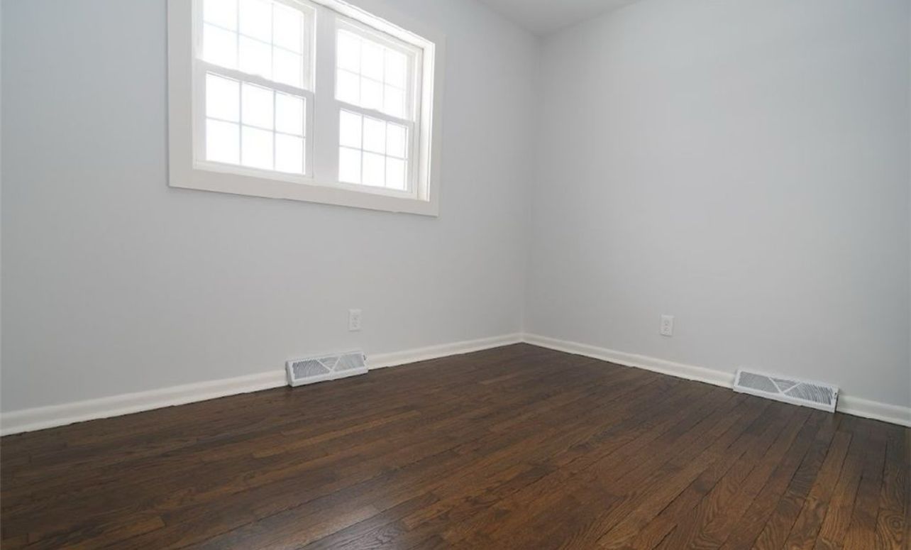 An empty room with light gray walls, dark wood floors, a white-trimmed window, and two floor vents.