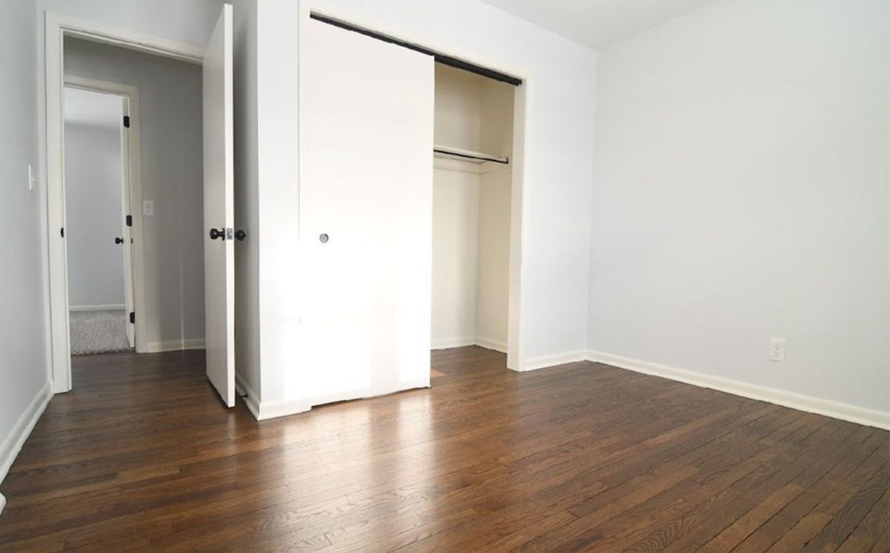 An empty room with dark hardwood floors, white walls, a sliding-door closet, and an open doorway leading to a hallway.