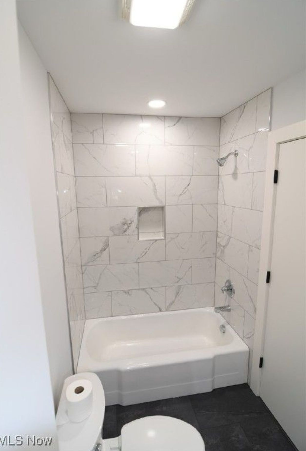 A white bathtub with marble-look tile walls, a built-in recessed niche, and a shower head, next to a white toilet.