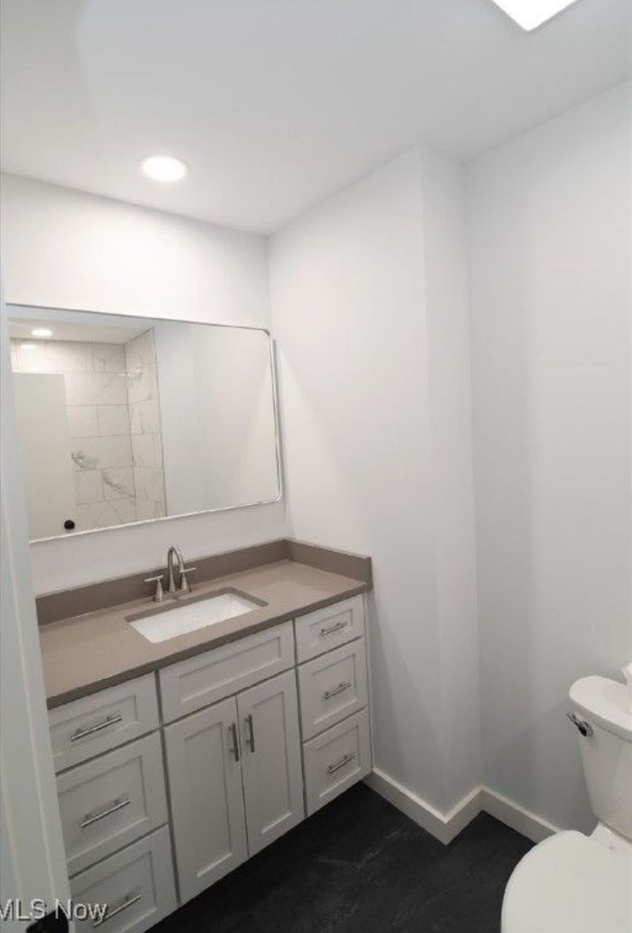 A modern bathroom with a light grey vanity, rectangular sink, large mirror, and white walls above dark flooring.