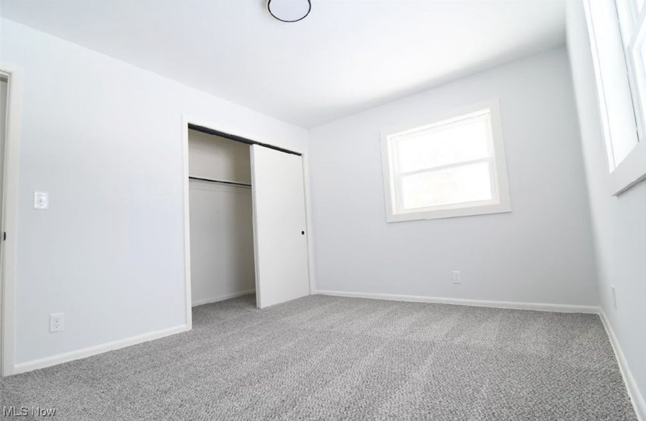 An empty bedroom with light gray walls, gray carpet, a closet with white sliding doors, and a window on the right.