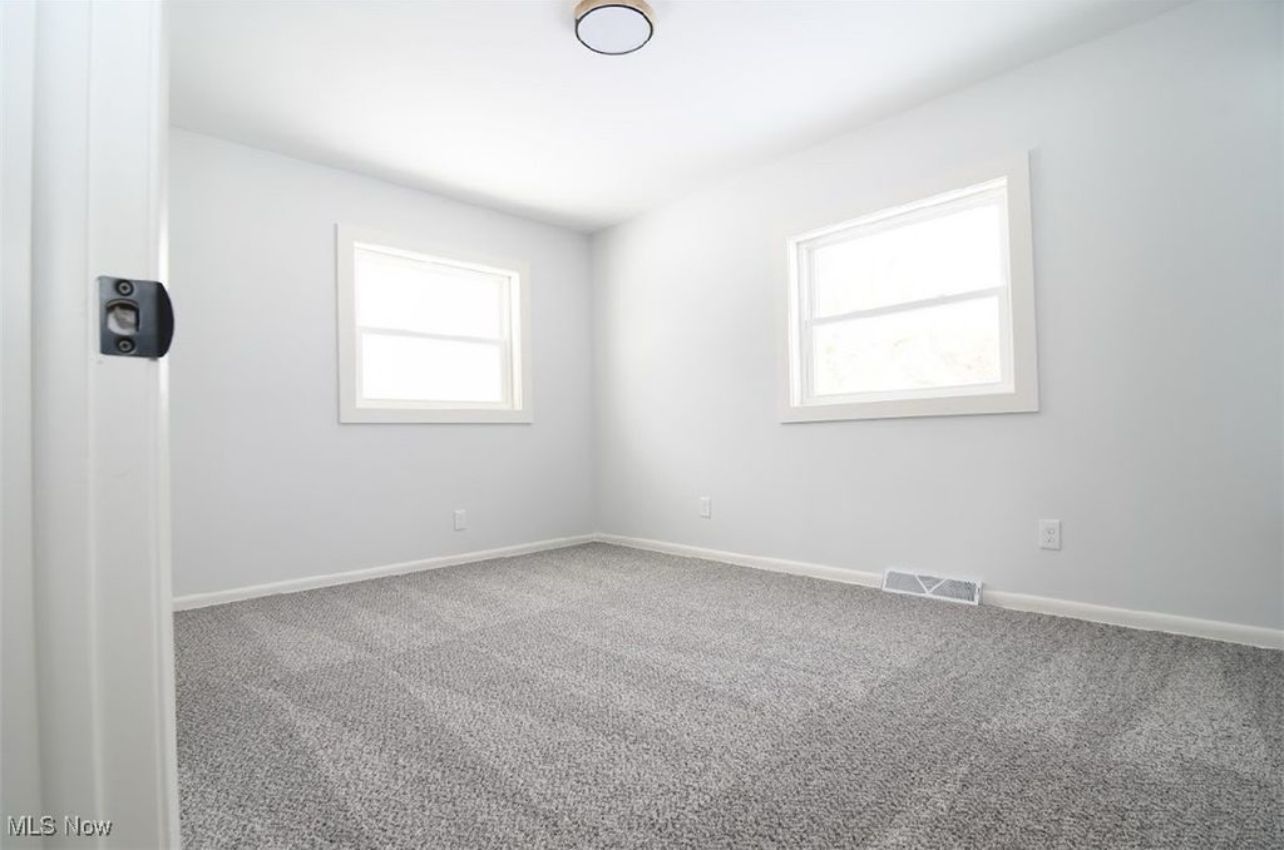 An empty bedroom with light gray walls, gray wall-to-wall carpeting, and two windows.