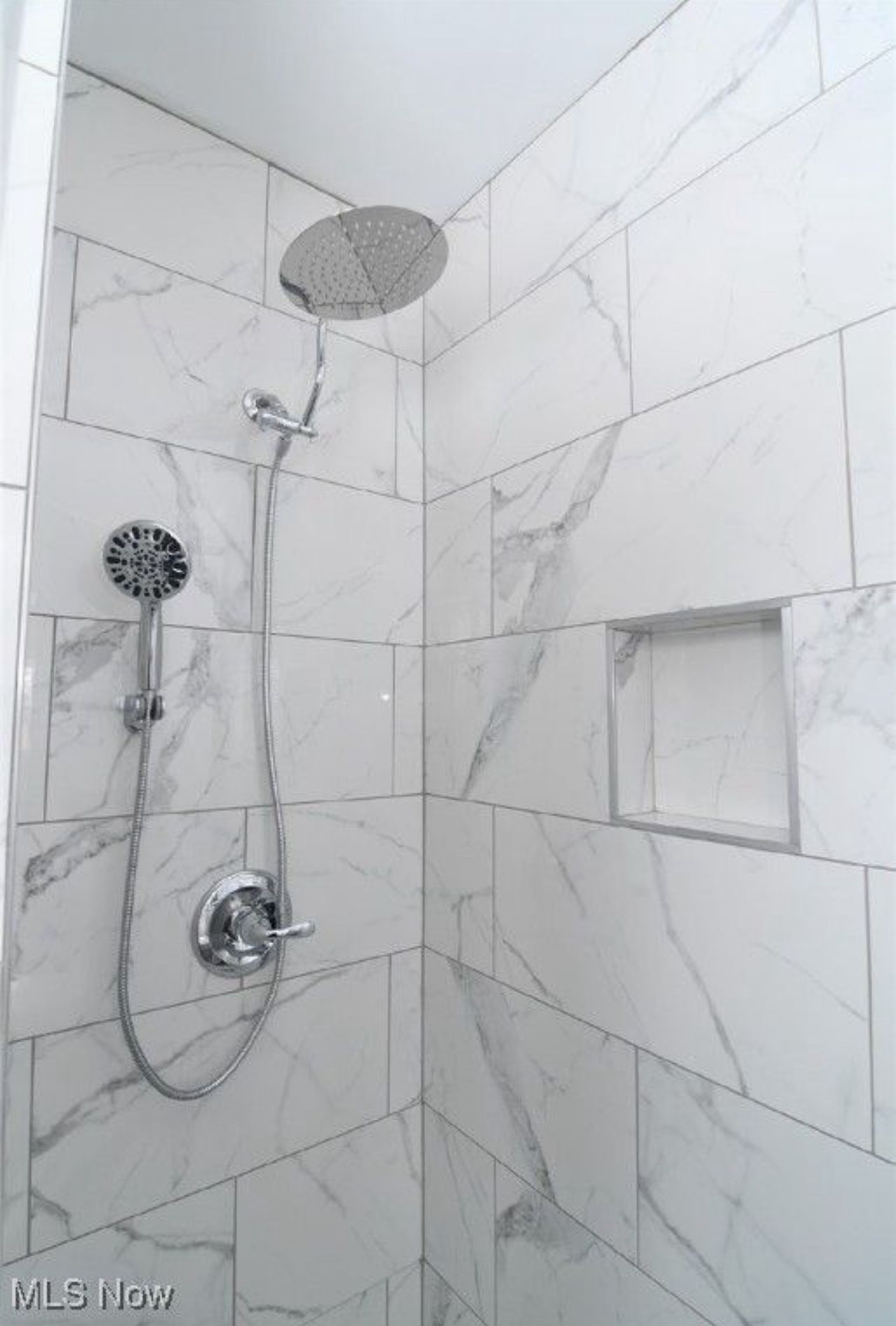 A tiled walk-in shower with white marble-patterned walls, a rainfall showerhead, a handheld sprayer, and a wall niche.