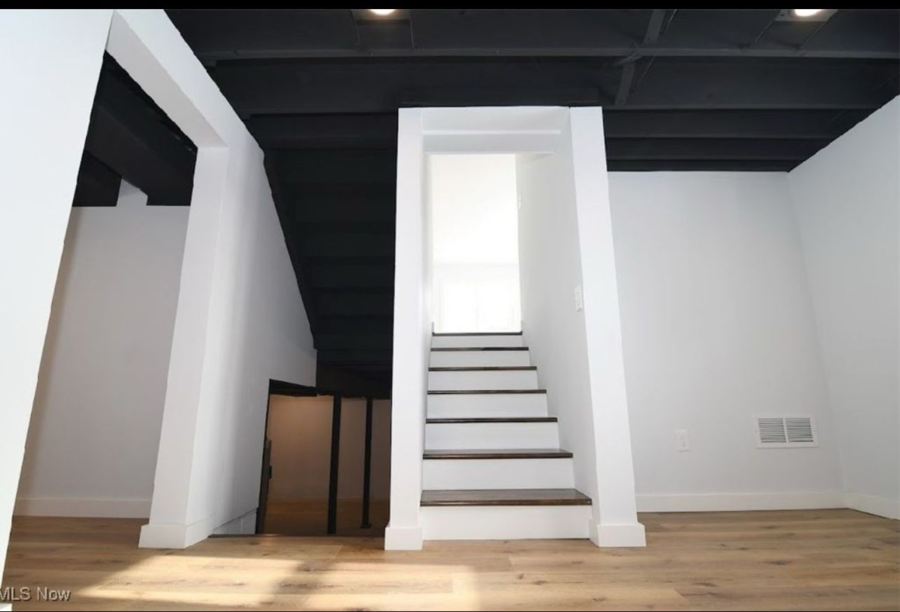 A modern room with white walls, light wood flooring, a black ceiling, and a central staircase leading upward.