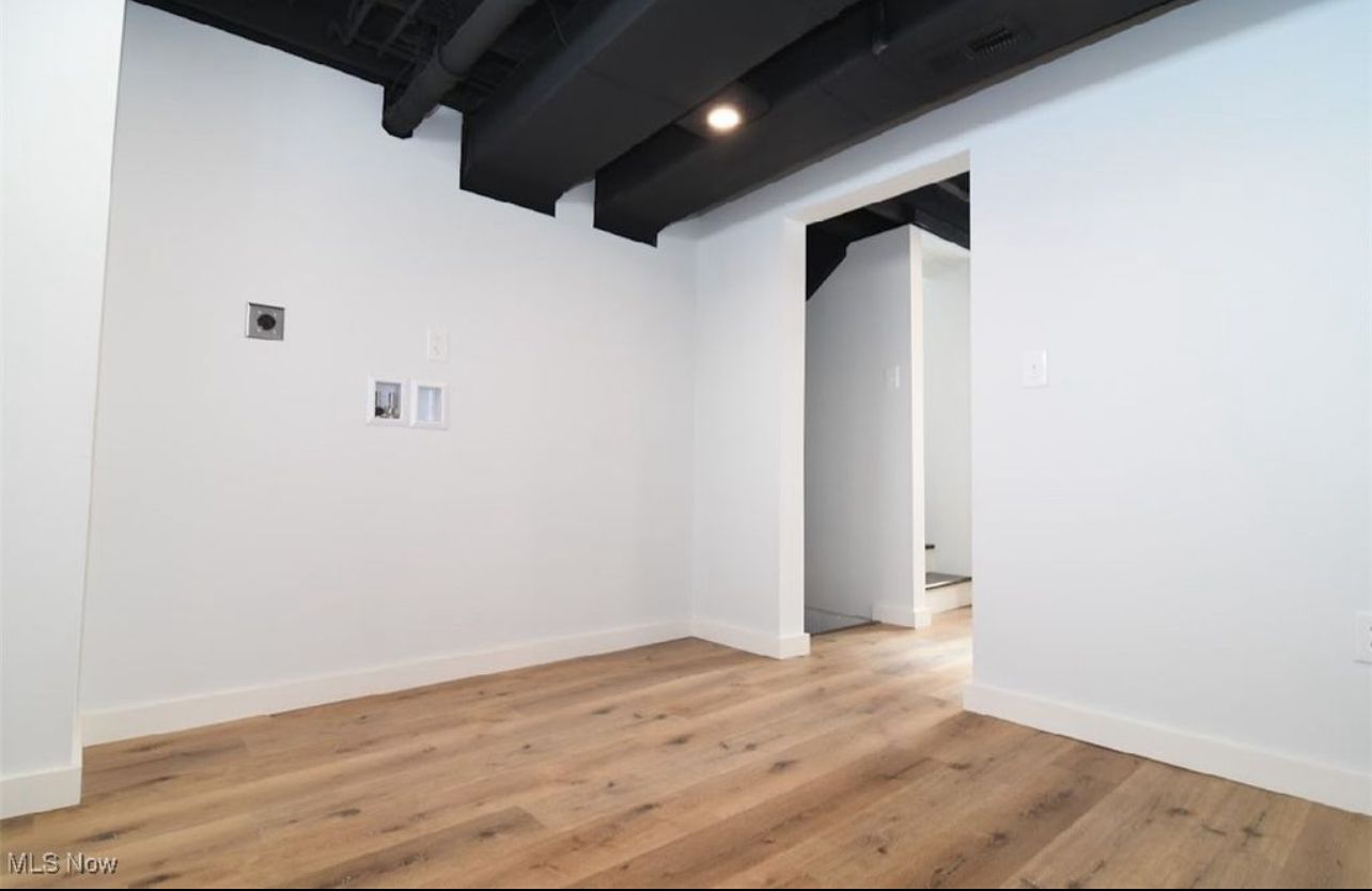 An unfurnished room with white walls, light wood floors, and black exposed ceiling beams, leading into a hallway.