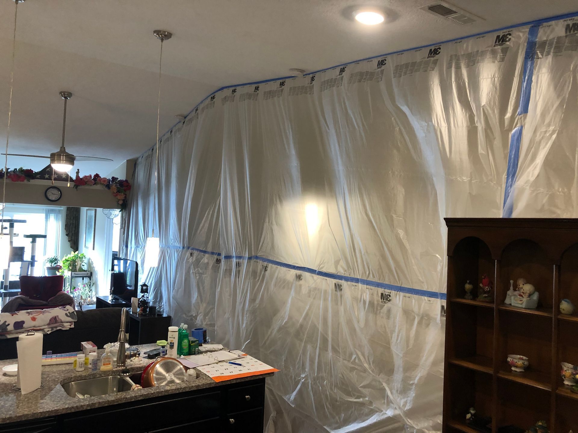 A kitchen wall covered with large sheets of clear plastic, secured with blue painter's tape, for a home renovation project.