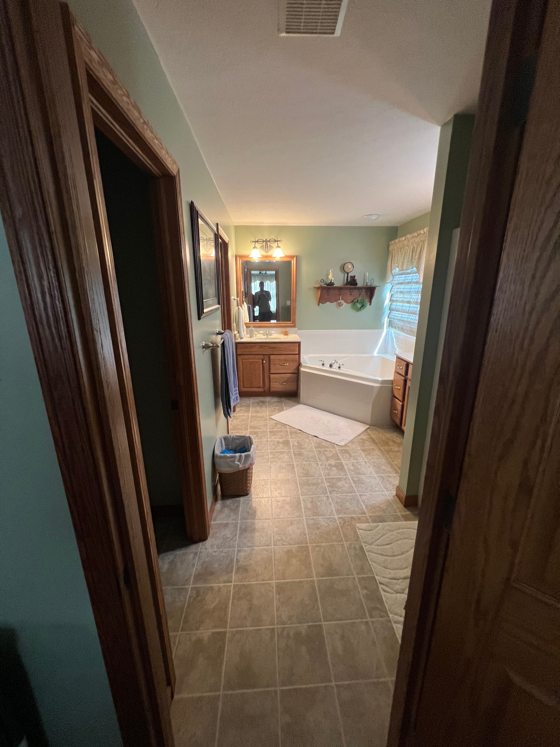 A doorway view into a light green bathroom with tan tile flooring, a wooden vanity with a mirror, and a soaking tub.