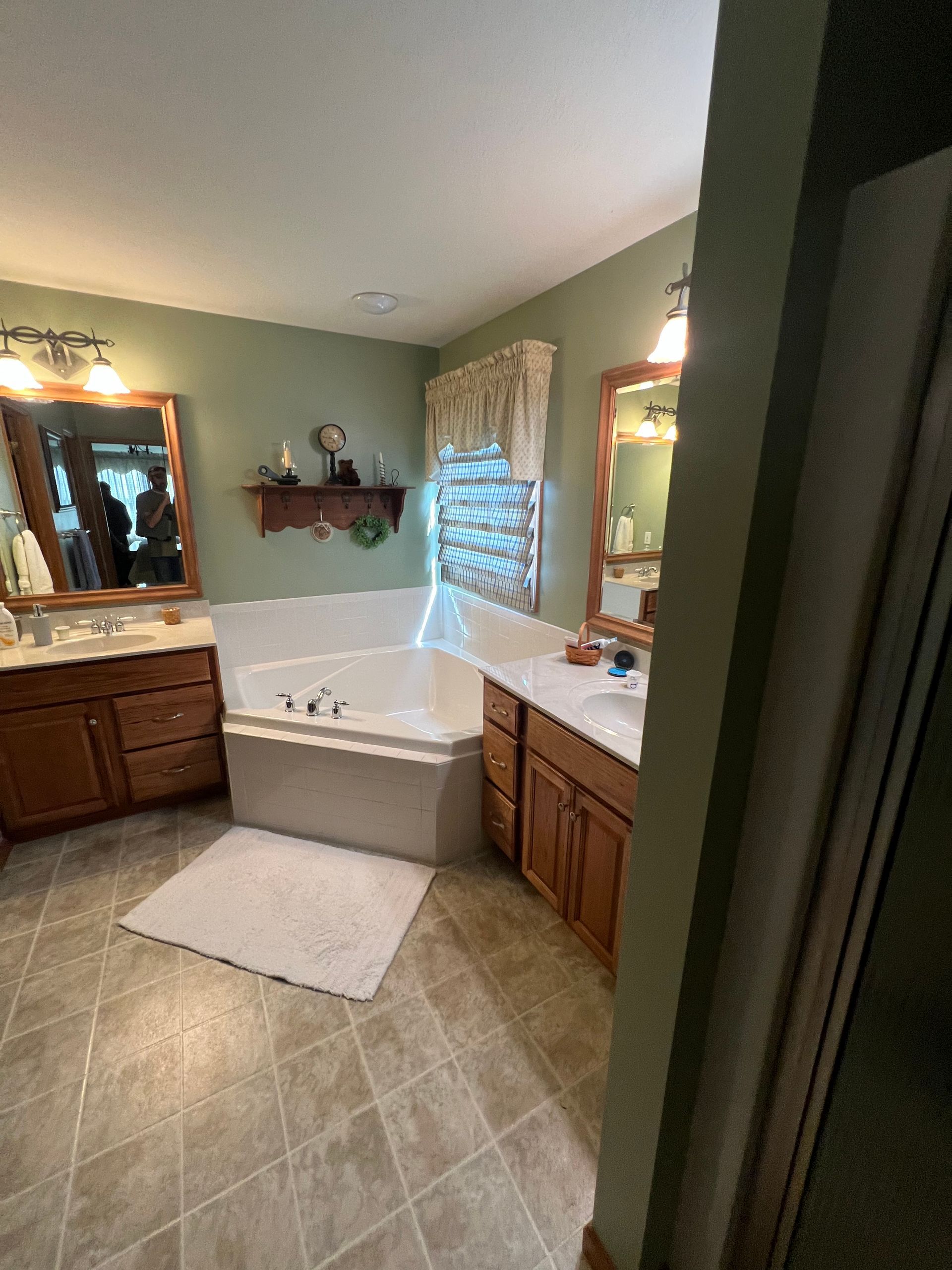 A bathroom with sage green walls, dual wooden vanities, a corner soaking tub, and tiled flooring.