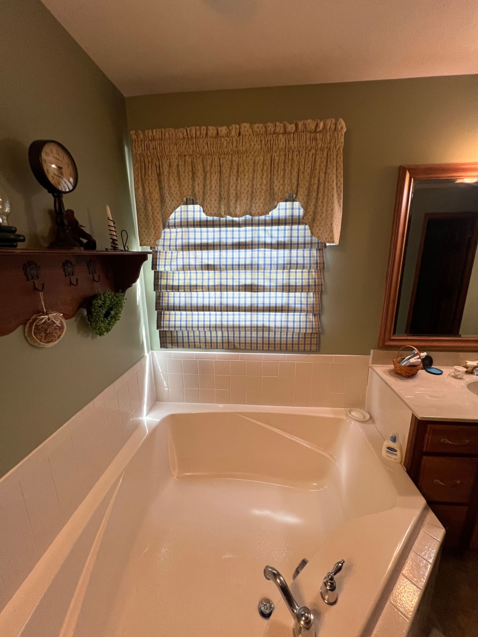 A bathroom corner featuring a large beige soaking tub under a window with a valance and brown woven shade, next to a sink.
