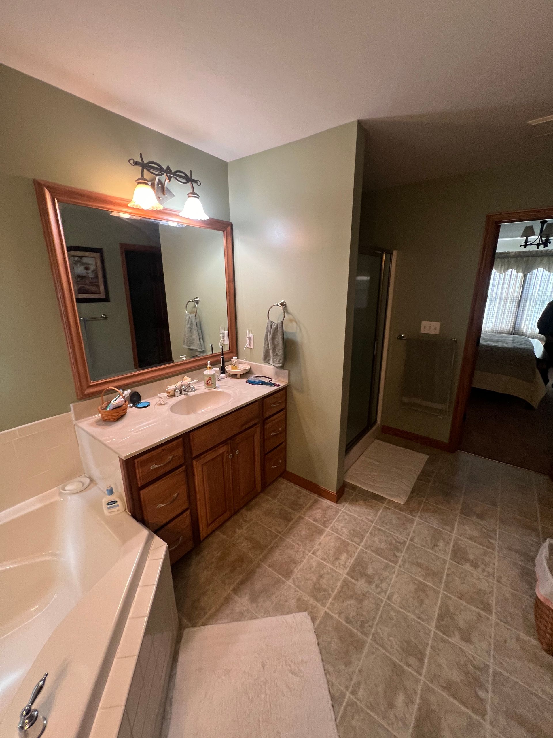 A bathroom with sage green walls, a wood-framed mirror over a wood vanity, and a tiled floor leading to a bedroom doorway.