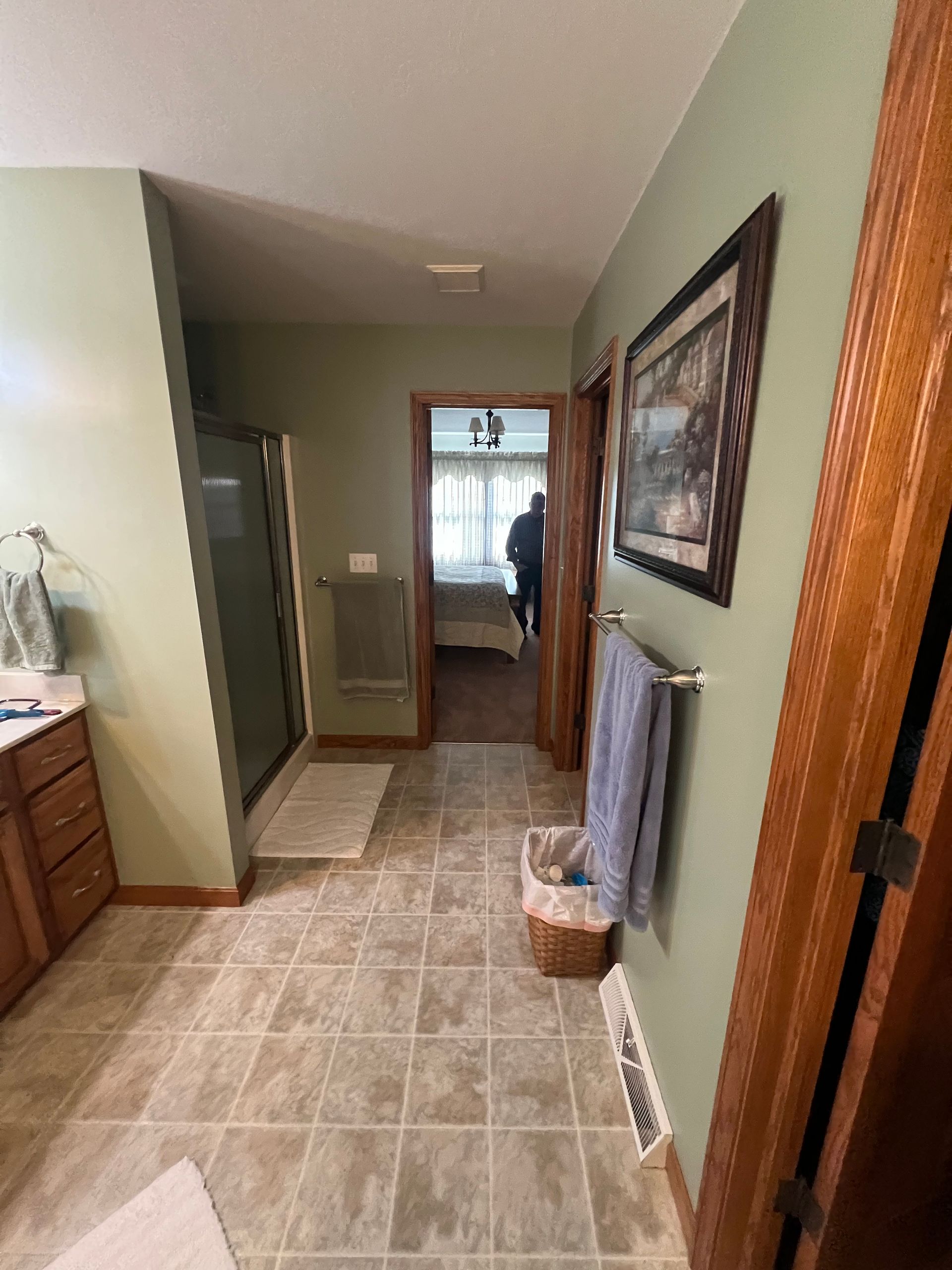 A view down a bathroom hallway with sage green walls, tiled floors, a shower, a towel rack, and a doorway to a bedroom.