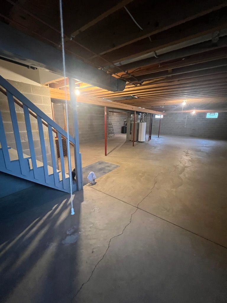 An unfinished basement with gray concrete walls, a wooden staircase, and an exposed ceiling structure.