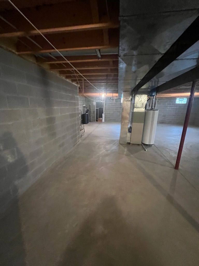 An unfinished basement with gray concrete block walls, a concrete floor, exposed wooden ceiling joists, and a water heater.