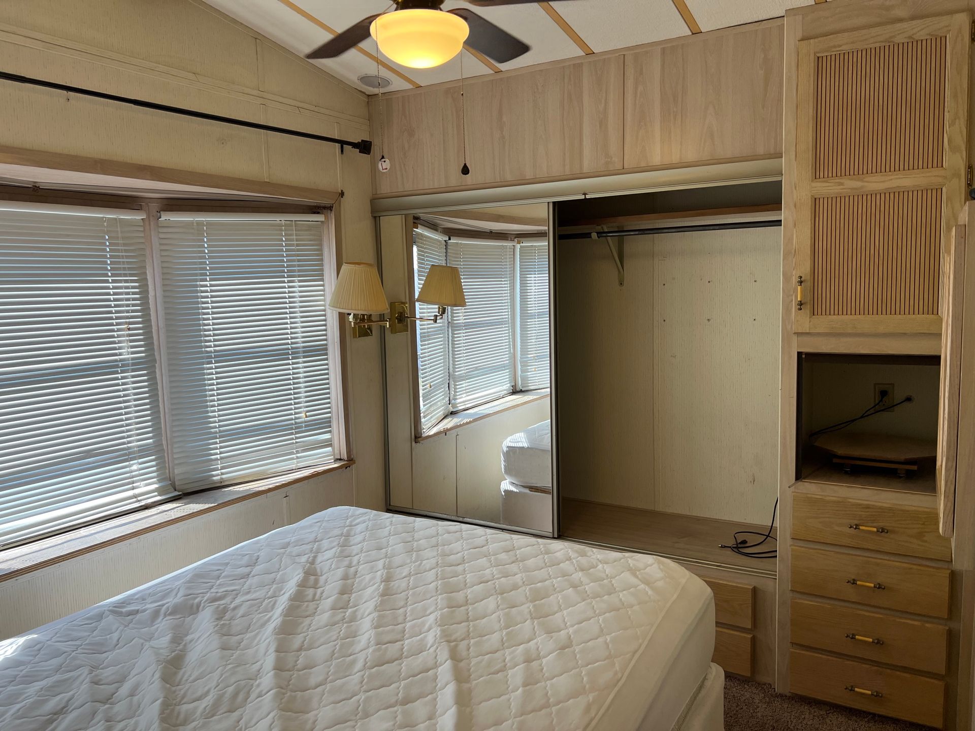 A bedroom with a bed, large window with blinds, mirror-doored closet, and wood cabinetry in a neutral color palette.