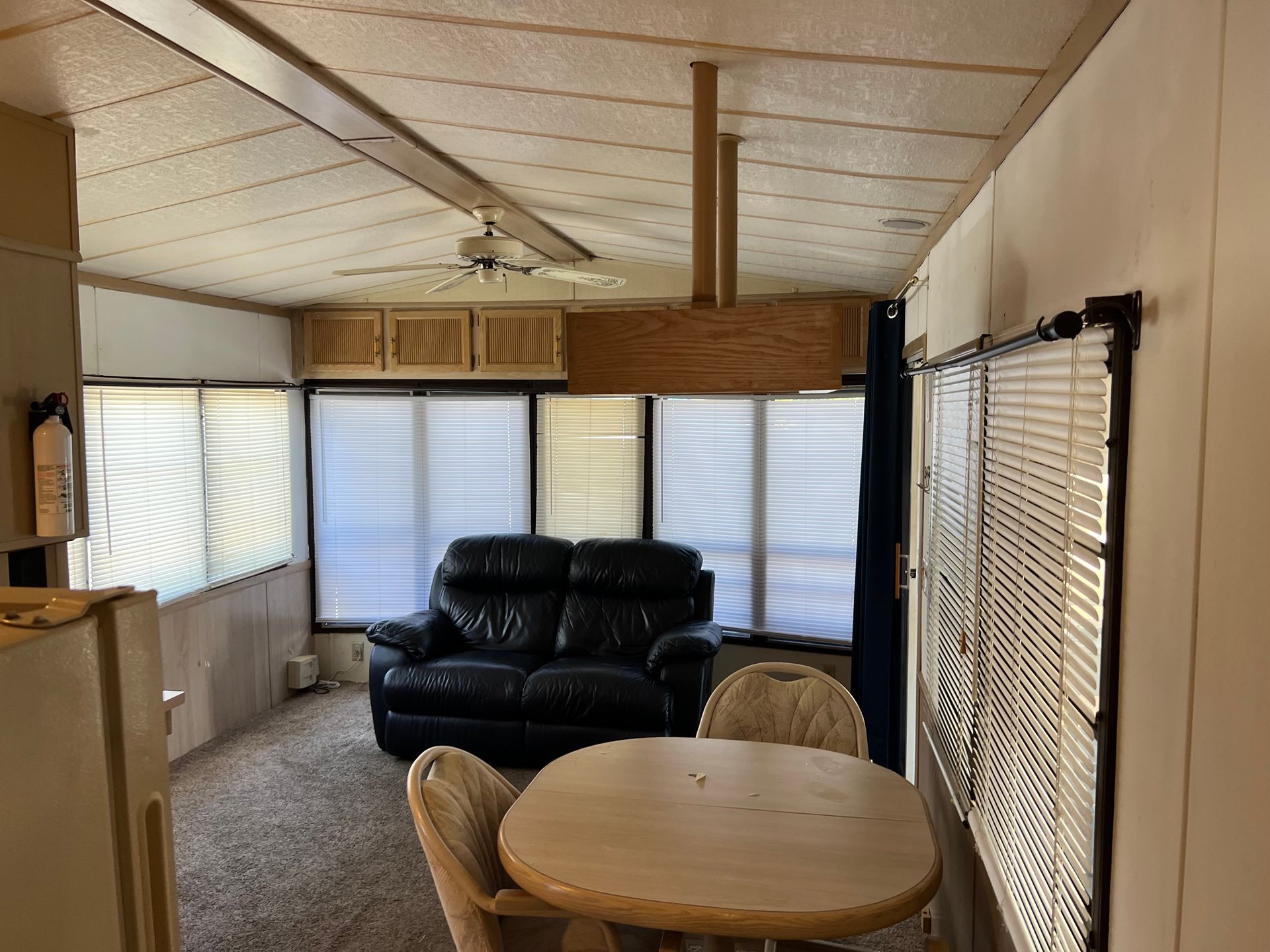 A living and dining area in a mobile home featuring a dark blue loveseat, a round light wood table, and window blinds.