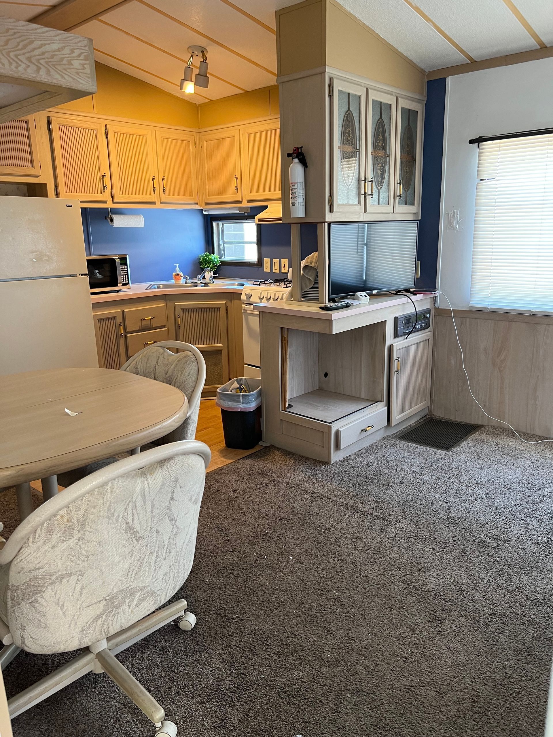 A kitchen and dining area with light wood cabinets, blue backsplash, a small round table, and textured carpet flooring.