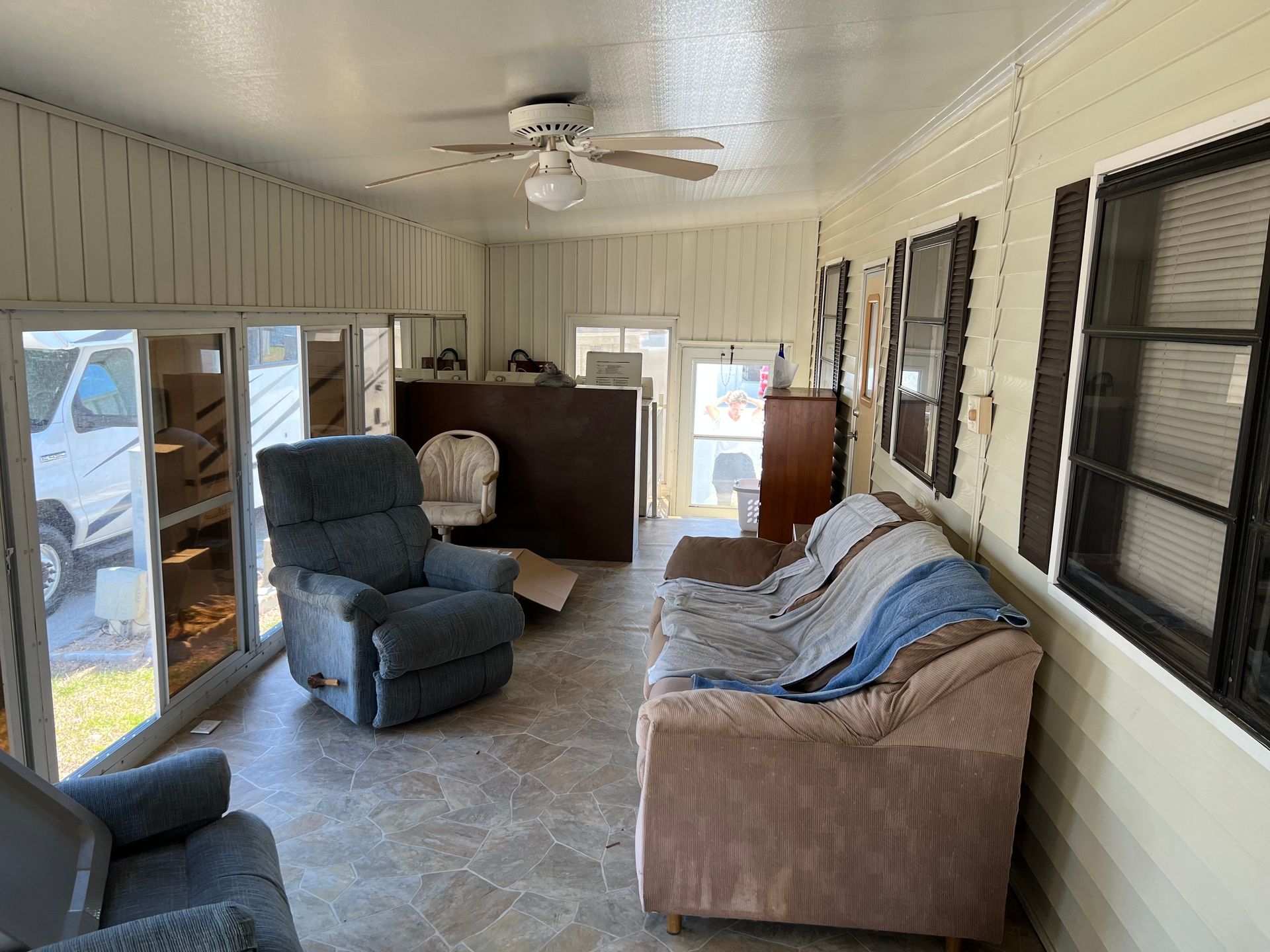 A sunroom with tile flooring, a blue recliner, a couch with blankets, and windowed walls looking out to a white vehicle.
