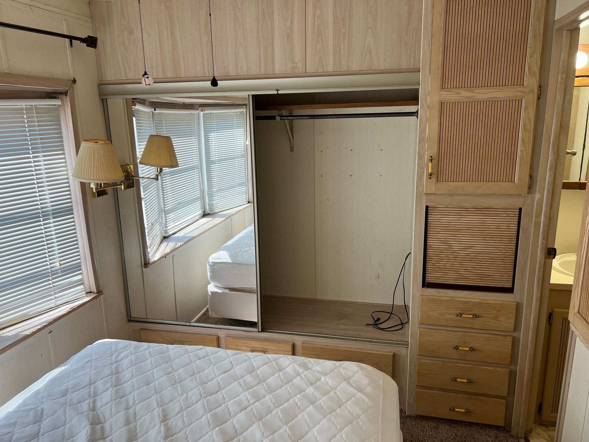 A bedroom in a camper with a quilted white bed, a large mirrored wardrobe, built-in wooden drawers, and a wall lamp.