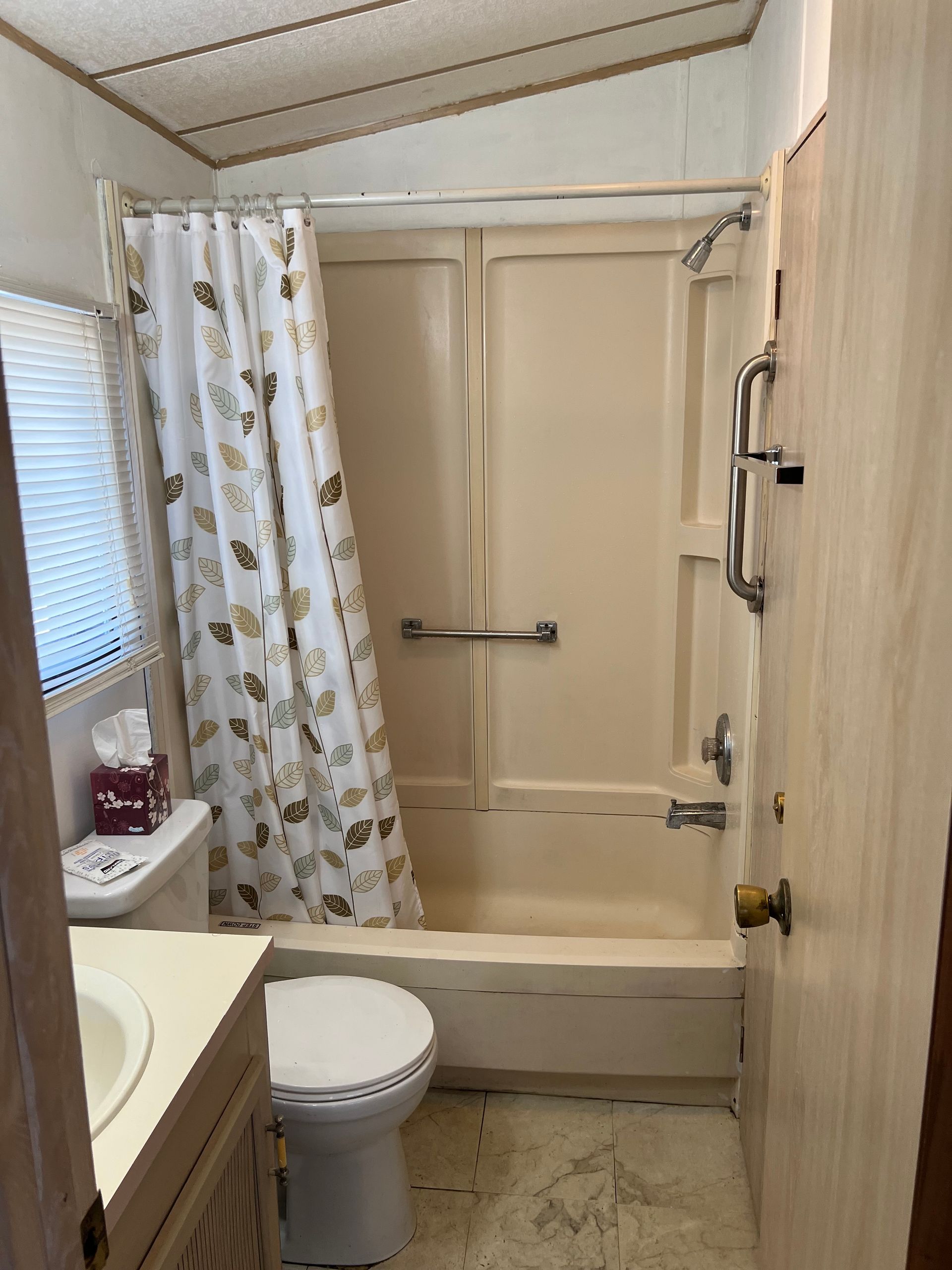 A beige bathroom with a tub-shower unit, a toilet, a sink vanity, and a white shower curtain with a leaf pattern.