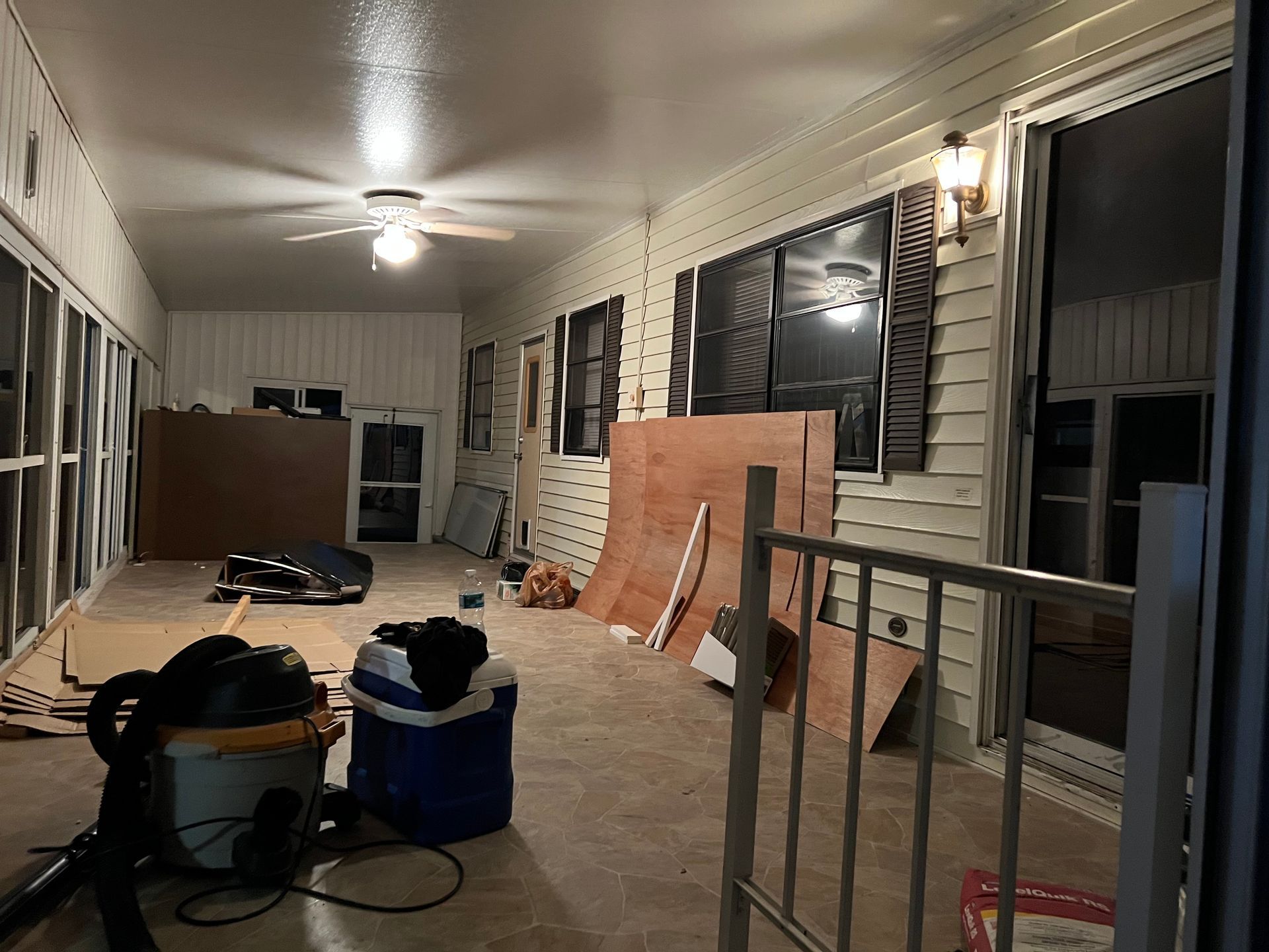A dimly lit, enclosed porch undergoing renovation with plywood sheets, a shop vacuum, and a cooler on the floor.