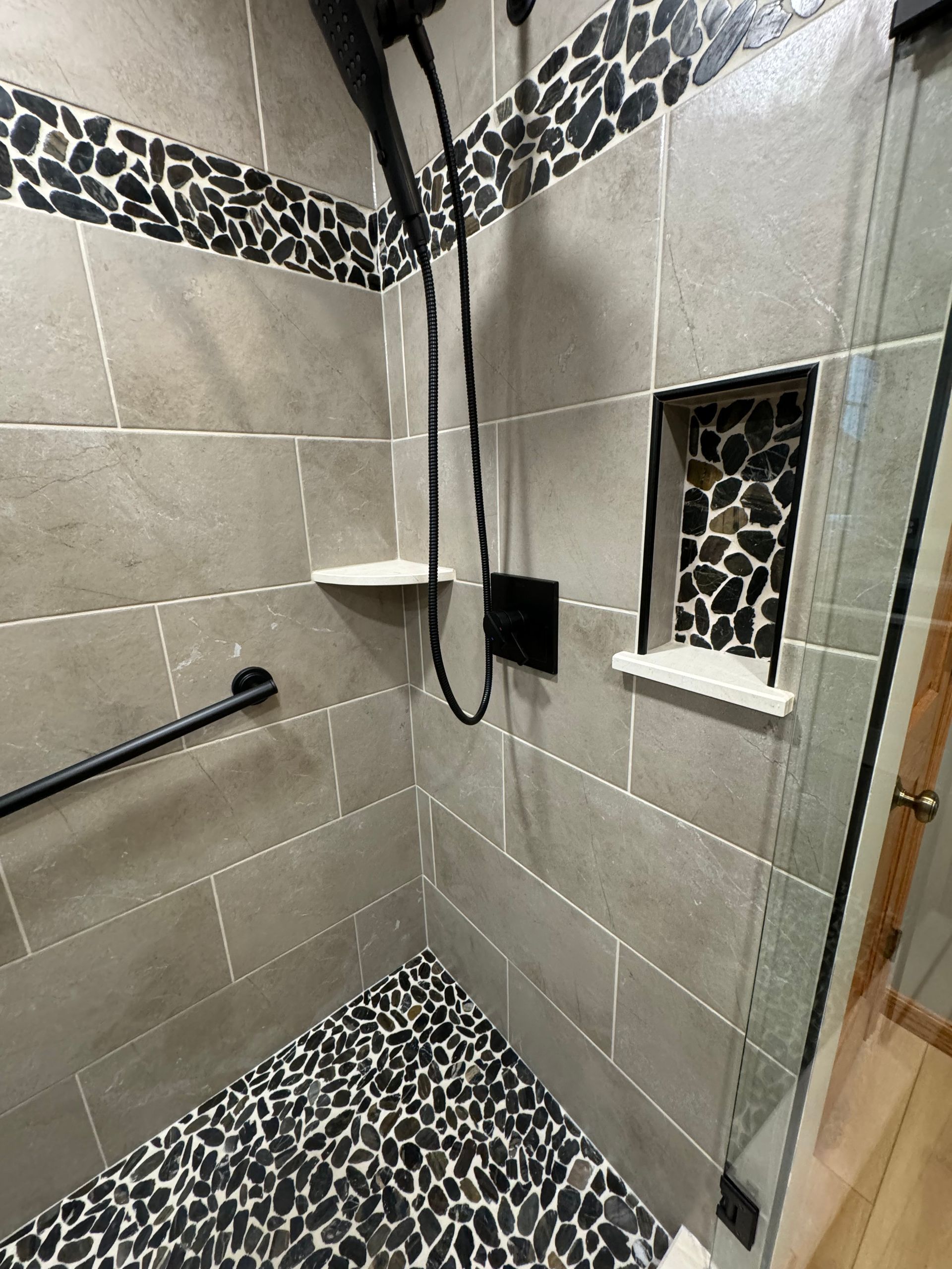 A walk-in shower with gray tiled walls, a black pebble floor, a built-in recessed shelf, and a black metal grab bar.