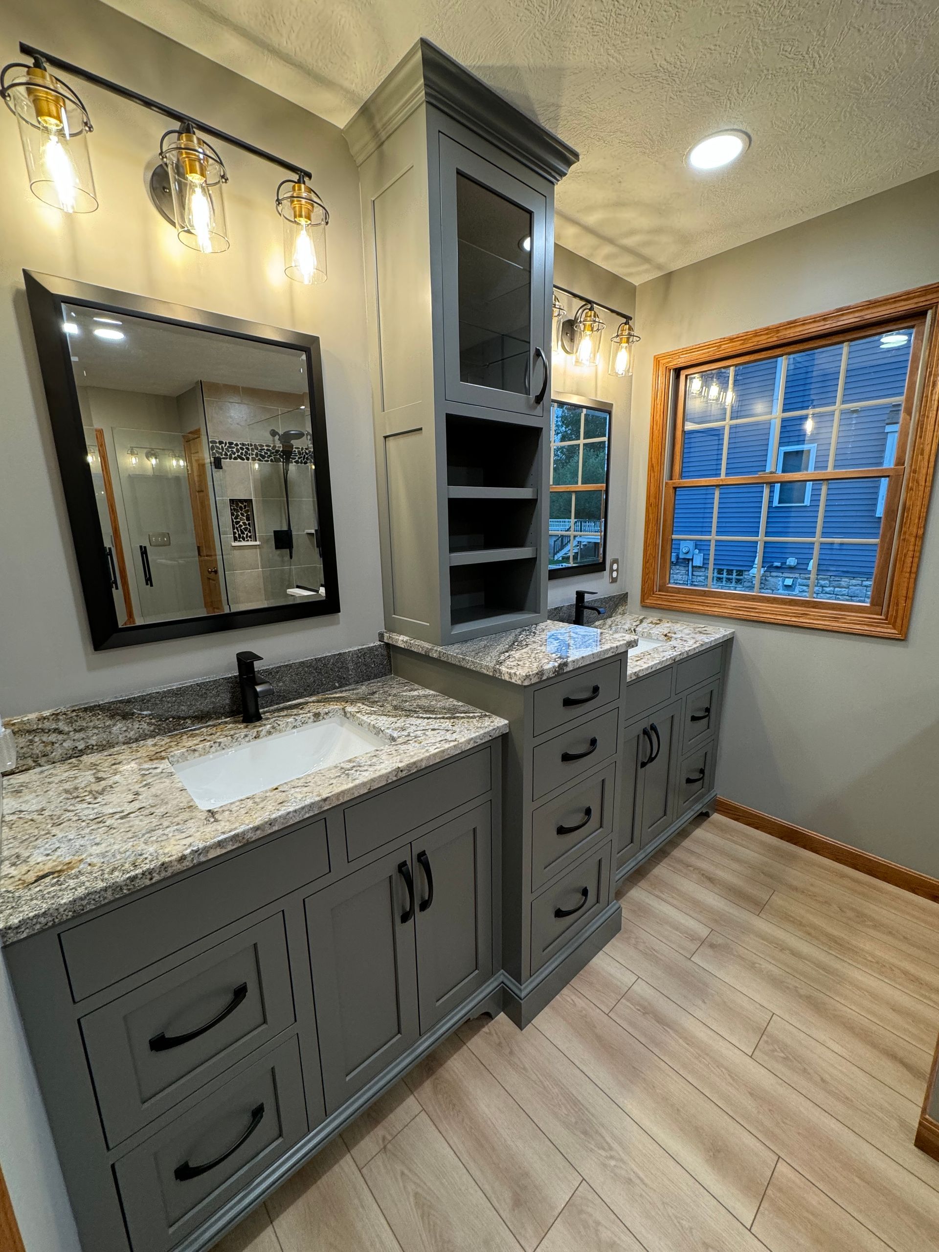 A bathroom vanity with gray cabinets, granite countertops, a tall storage hutch, and a wall-mounted mirror.