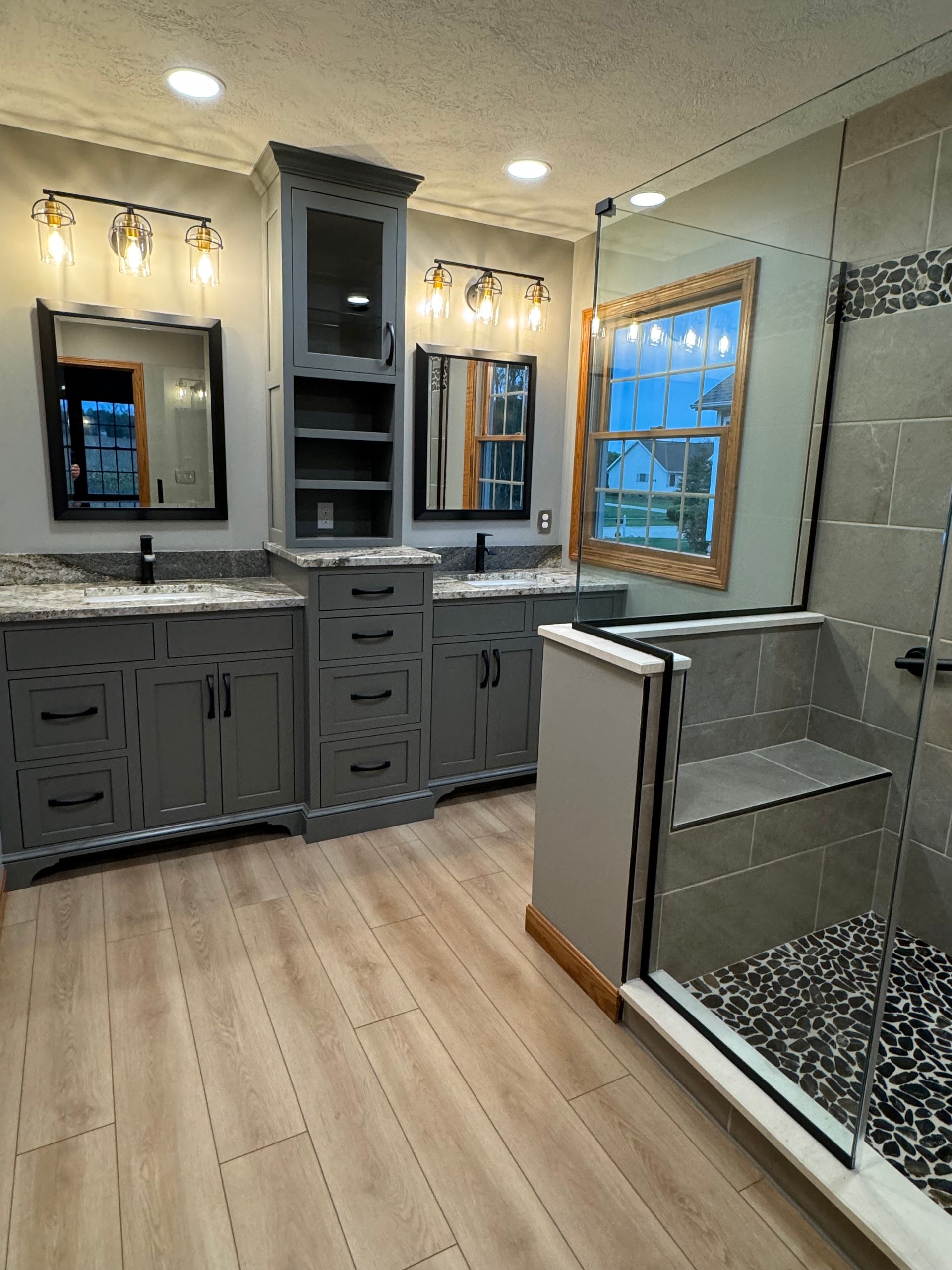 Modern bathroom with a gray double vanity, tower cabinet, two mirrors, and a glass-enclosed shower with tiled walls.