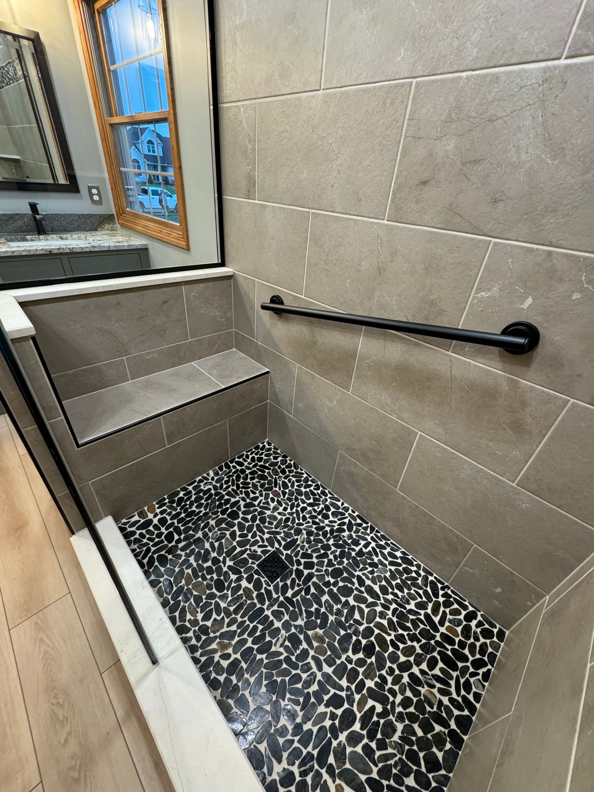 A walk-in shower with large beige tiles, a black pebble floor, a built-in stone bench, and a black horizontal grab bar.