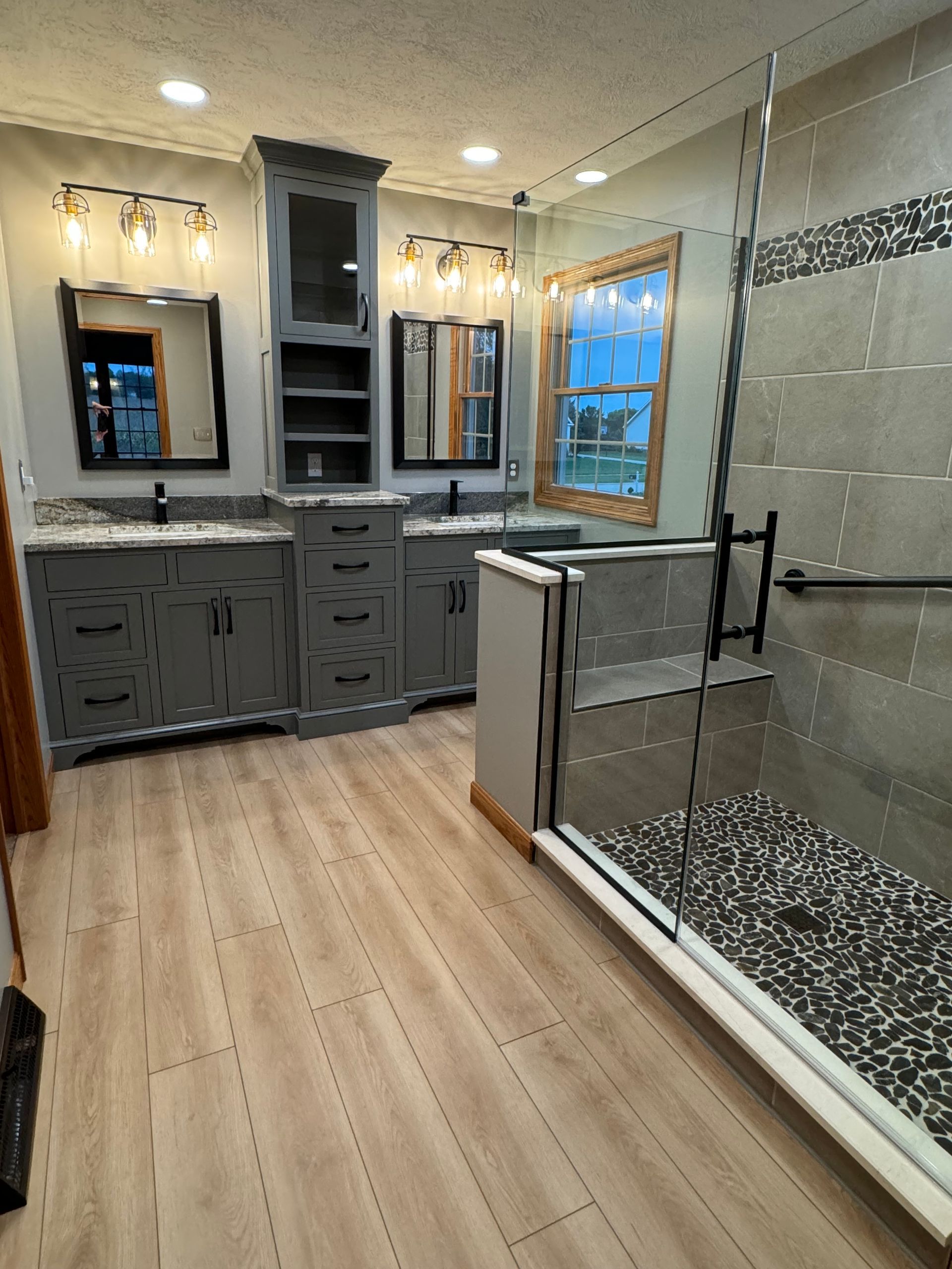 A bathroom featuring dual grey vanities, a central storage tower, wood-look flooring, and a walk-in glass shower.