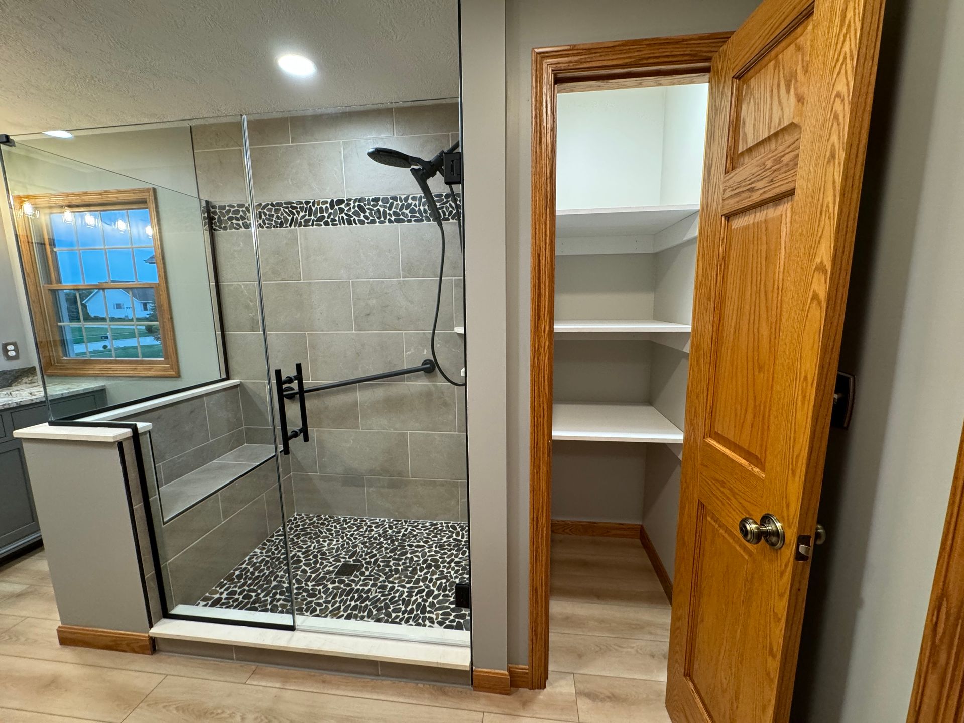 A bathroom with a tiled walk-in shower featuring a built-in bench and a small adjacent closet with white shelving.