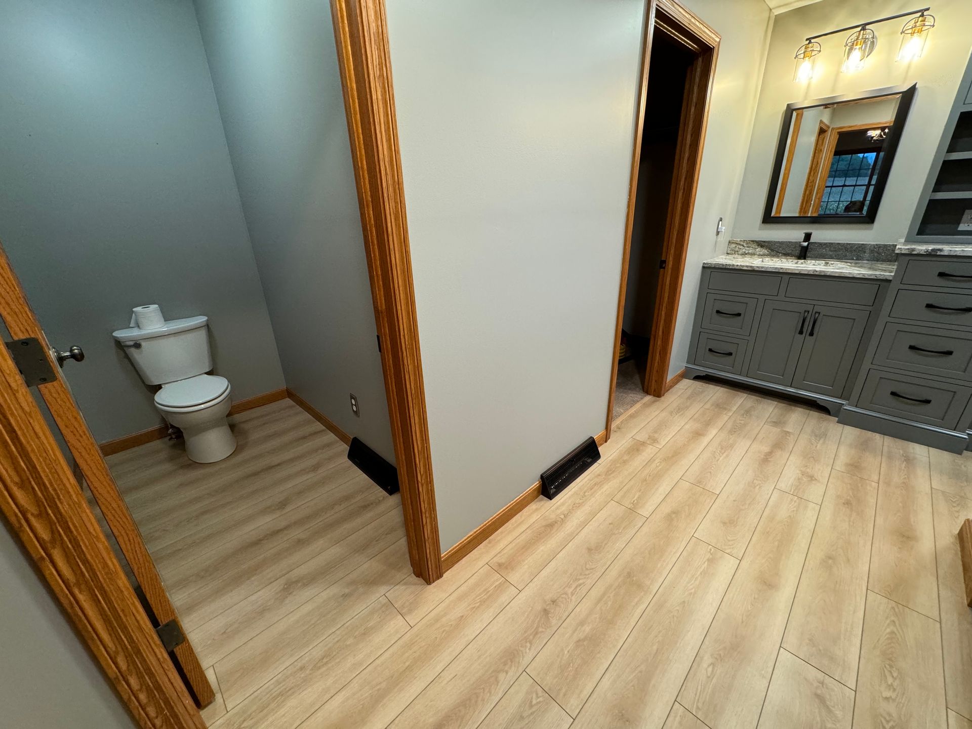 A bathroom featuring light wood-look flooring, a separate toilet area, and a grey vanity with a framed mirror and lights.
