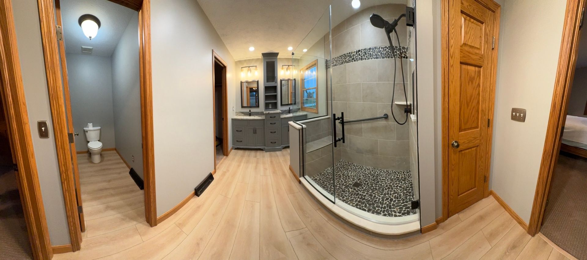 A panoramic view of a modern bathroom with wood-look floors, a glass-enclosed shower, a double vanity, and a private toilet.