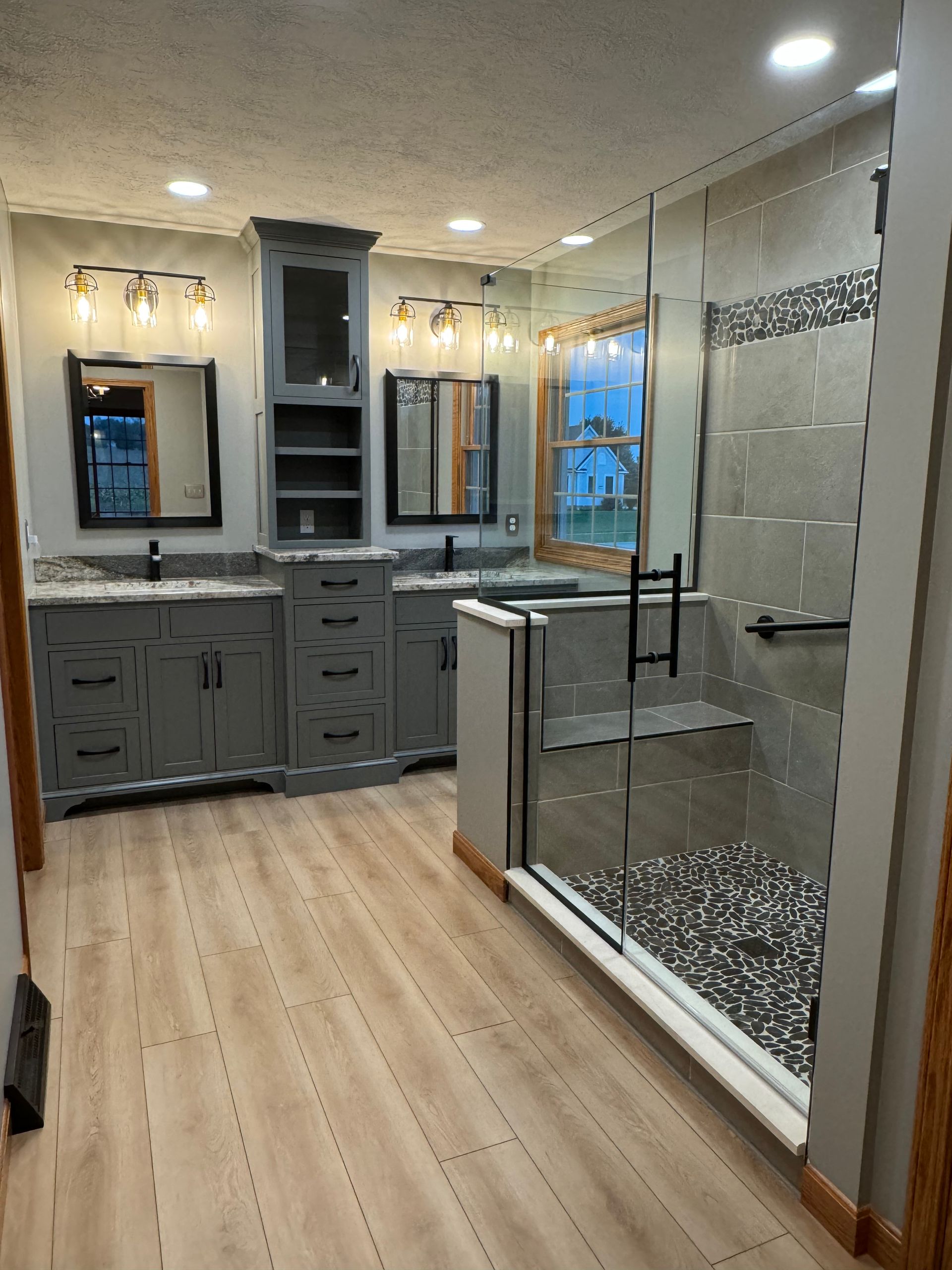 A modern bathroom featuring gray cabinets, a double vanity, a glass-enclosed shower with pebble floor, and light wood floors.