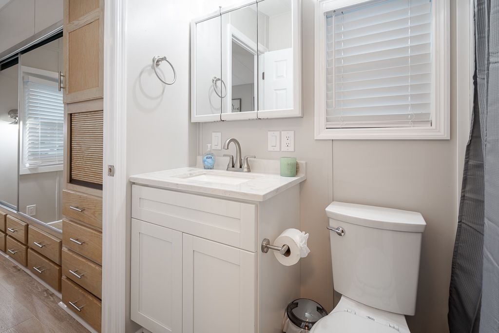 A bright bathroom features a white vanity sink, a medicine cabinet, a toilet, and light-toned wooden storage drawers.