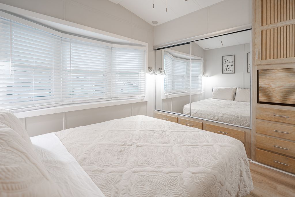 A bright, modern bedroom featuring a bed with a white quilt, large windows with white blinds, and mirrored closet doors.