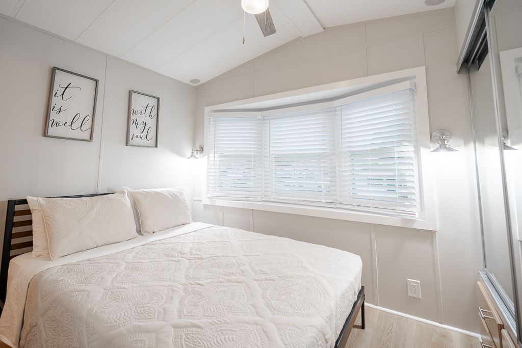 A brightly lit, modern bedroom featuring a bed with a white quilt, light gray walls, two framed prints, and a bay window.