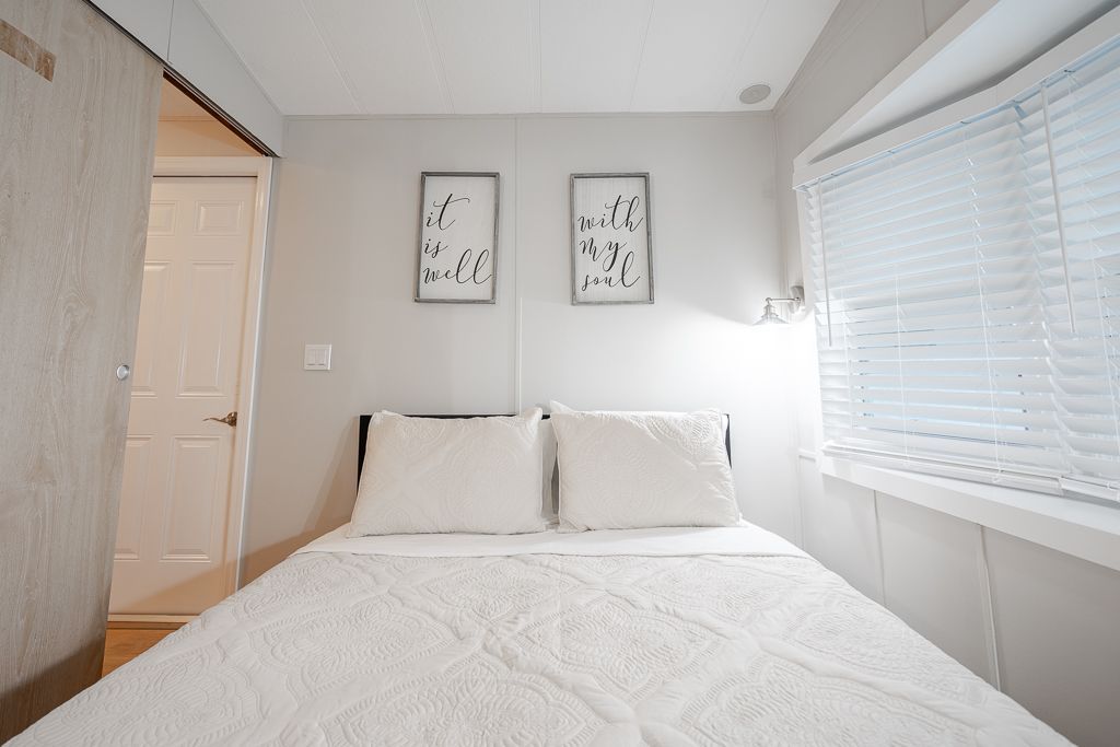 A bright, neutral-toned bedroom with a white bed, two framed wall pictures, and a window with white blinds.