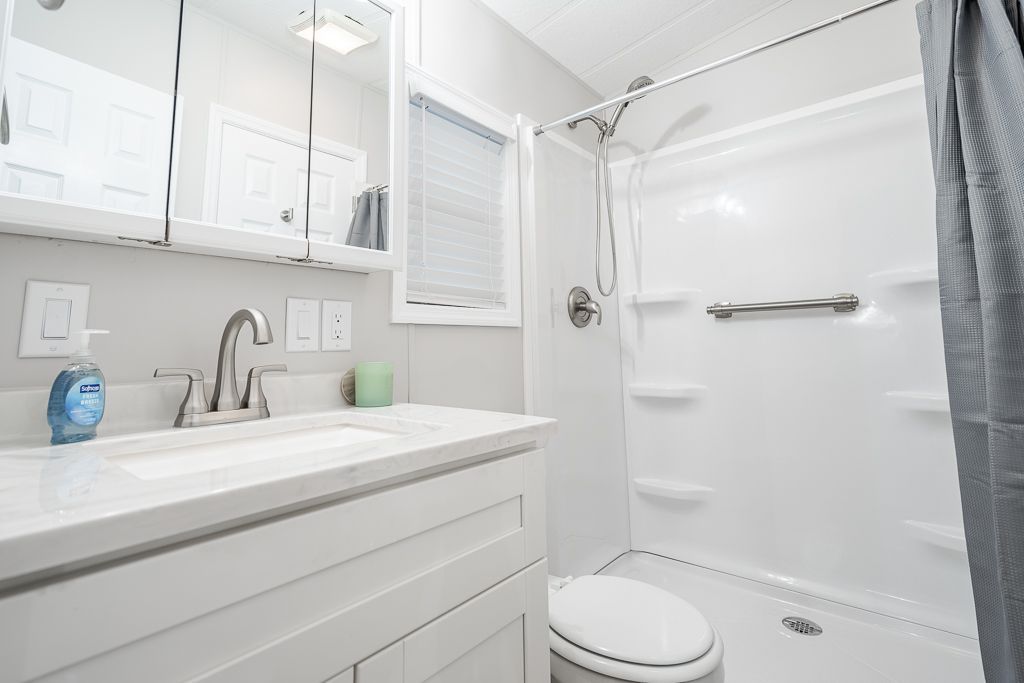 A modern, clean bathroom with a white vanity sink, a mirrored cabinet, a toilet, and a walk-in shower with a grab bar.