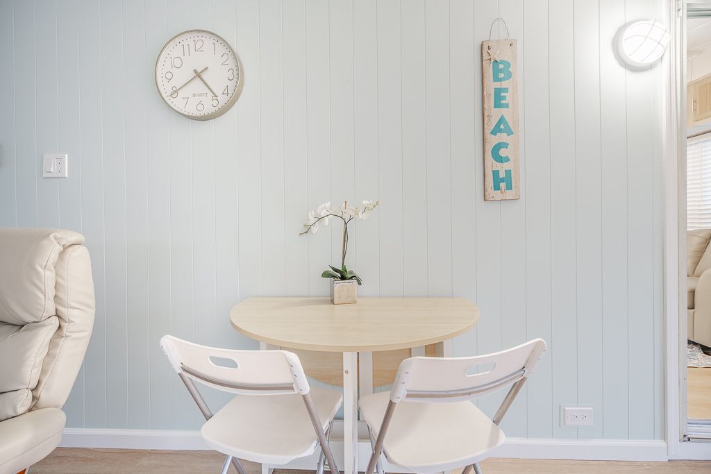 A small, light wood dining table with two white chairs, a potted orchid, a wall clock, and a 