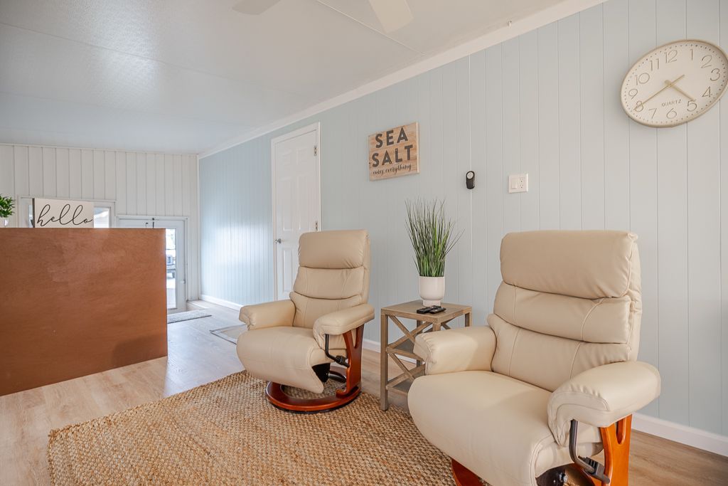 Two beige leather armchairs face a jute rug in a room with light blue wood-paneled walls, a wall clock, and decor.